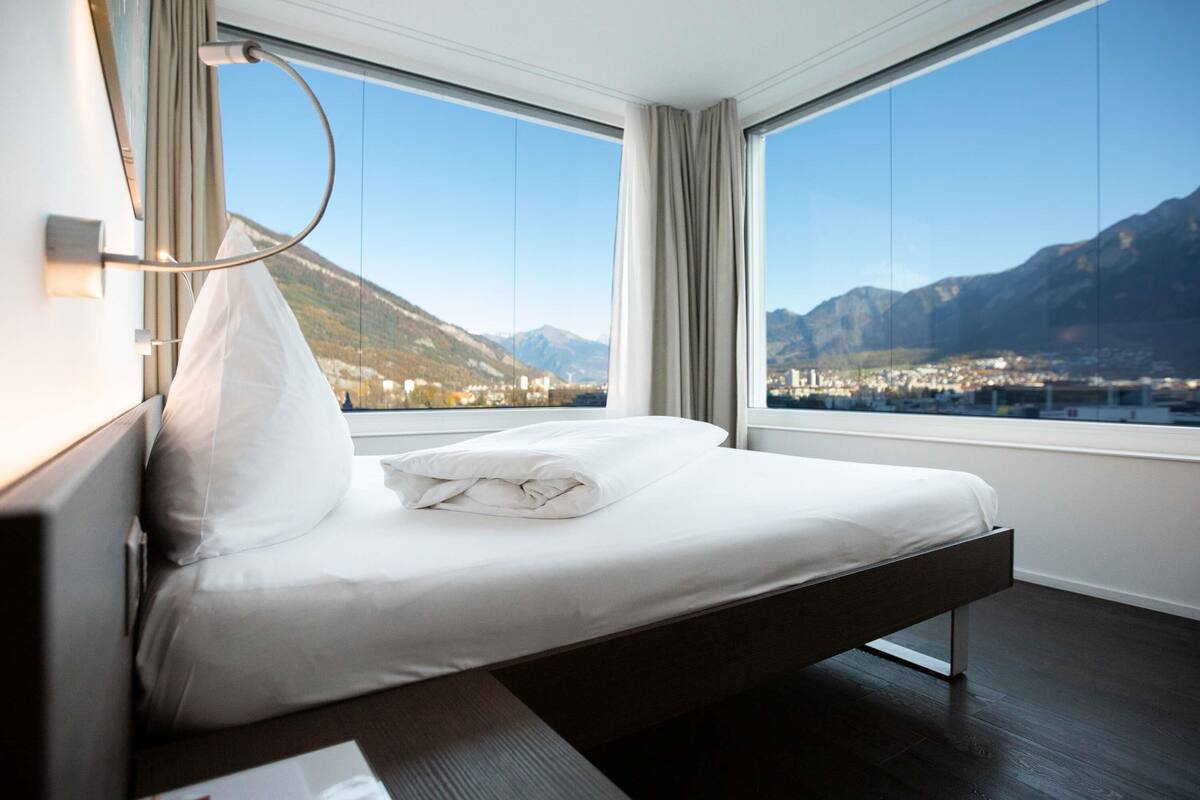 A modern hotel room with a bed, large windows, and a view of mountains.