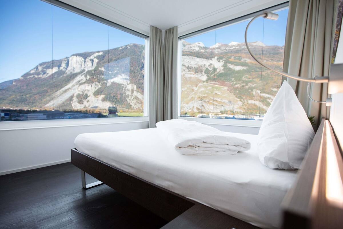 A hotel room with a bed, a view of mountains, and a large window.