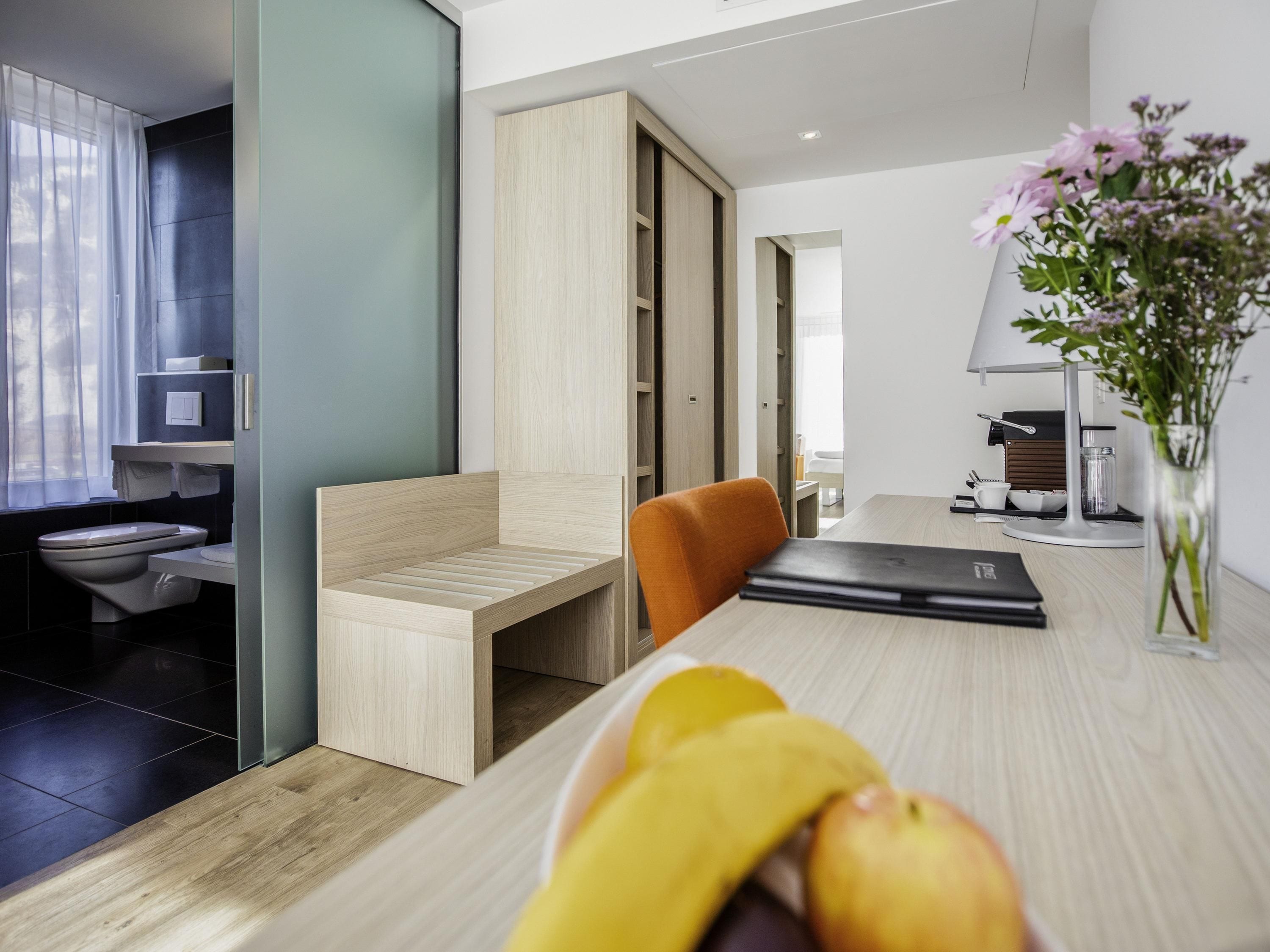 Ein modernes Zimmer mit Schreibtisch aus Holz, einem Stuhl, einem Laptop, einer Vase mit Blumen und Blick ins Badezimmer.