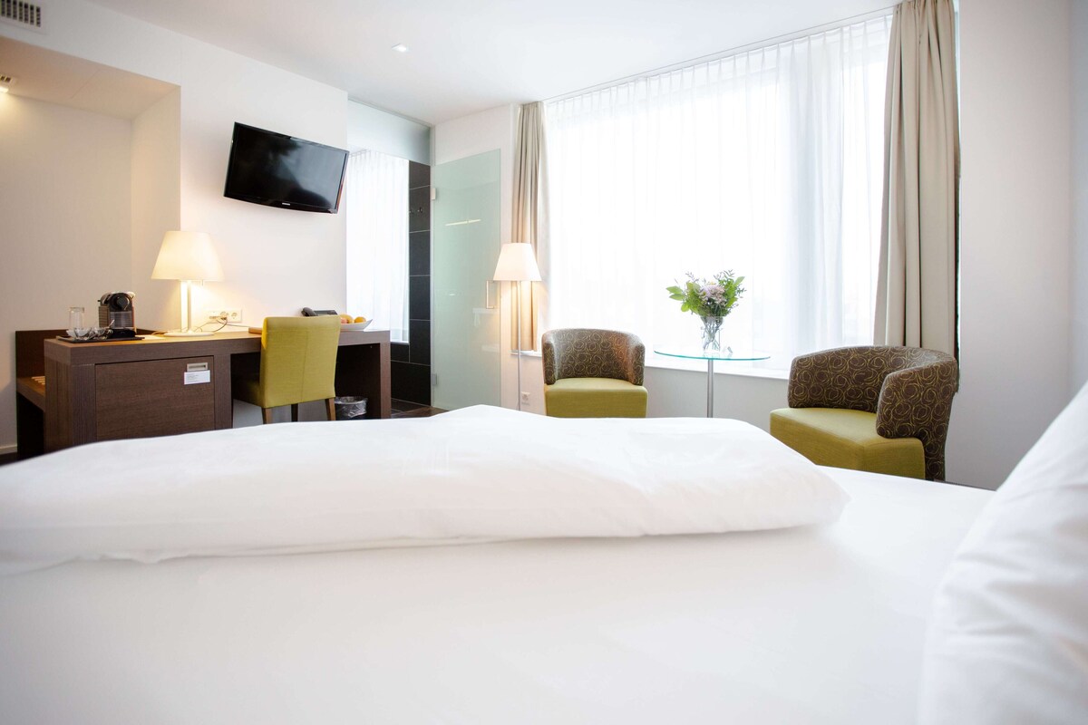 A hotel room with a bed, a desk, a chair, a TV, and a window with curtains.