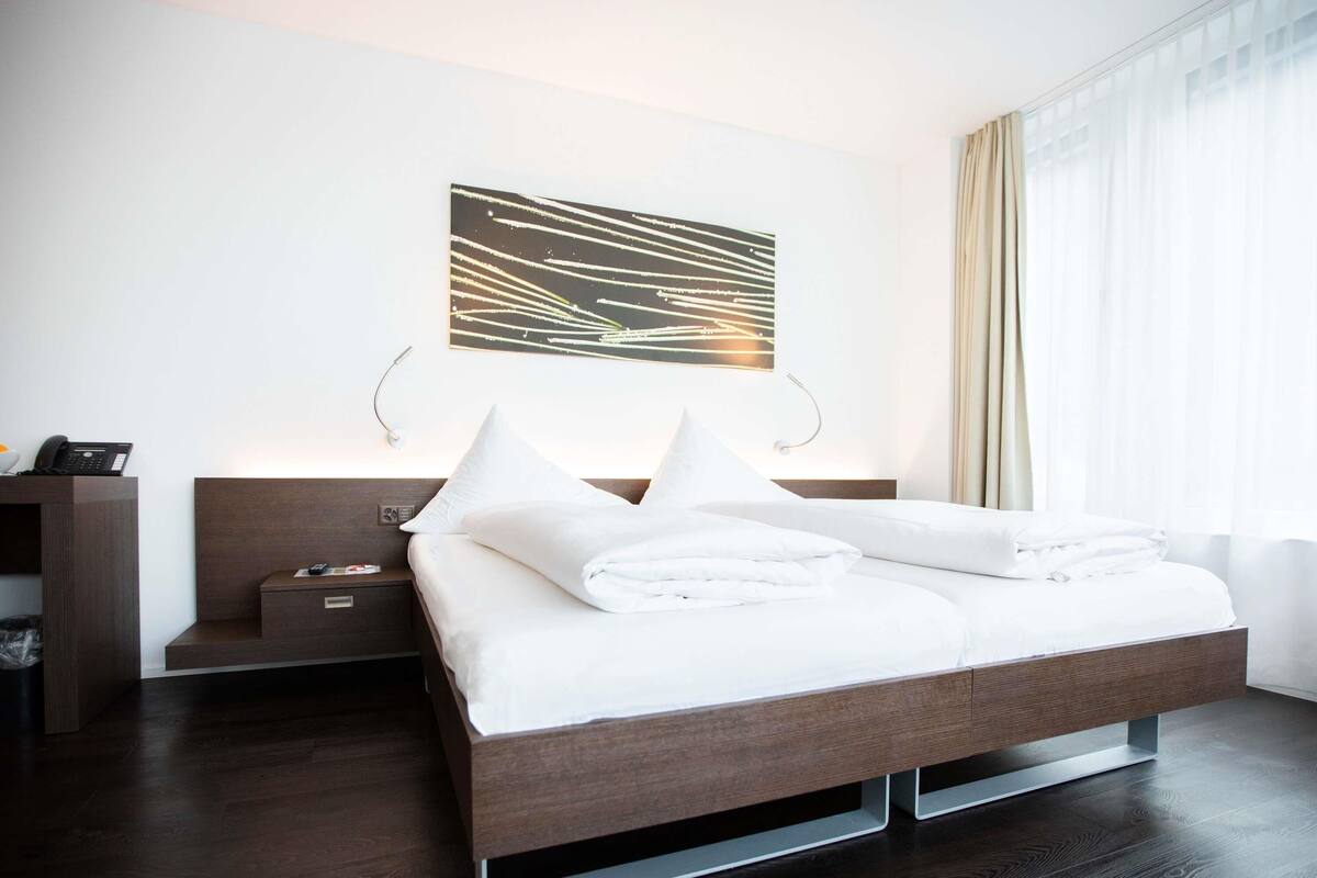 A modern hotel room with a large bed, bedside tables, a desk, and a wall-mounted artwork.