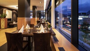 A modern dining area with tables set for two, large windows offering a city view, and a stone accent wall. - Mercure Chur City West (Chur)