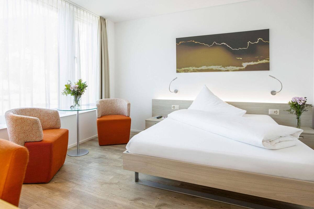 A hotel room with a bed, two chairs, a nightstand, a wall-mounted lamp, and a painting.