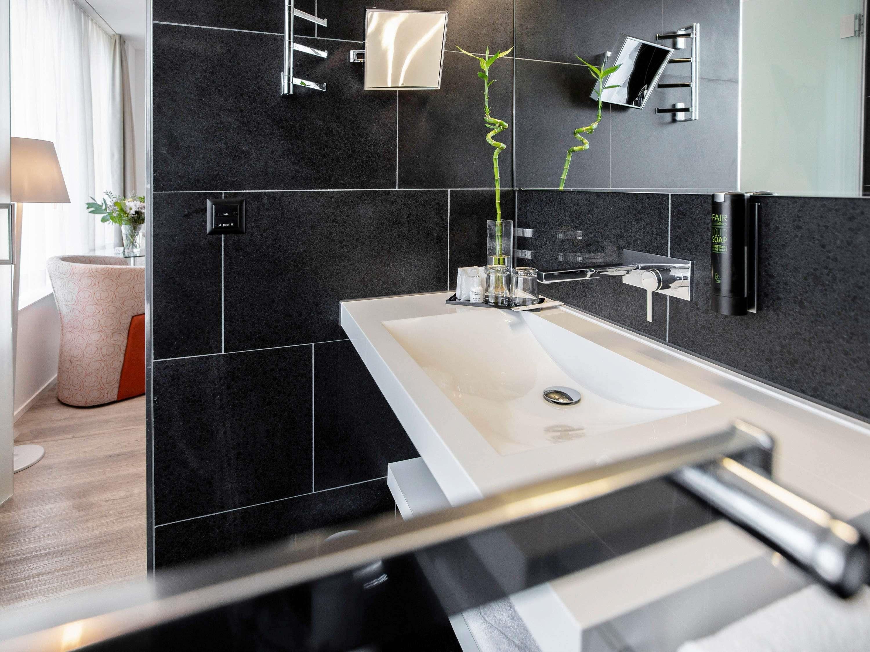 A modern bathroom with a white sink, black tiles, a mirror, and a plant.