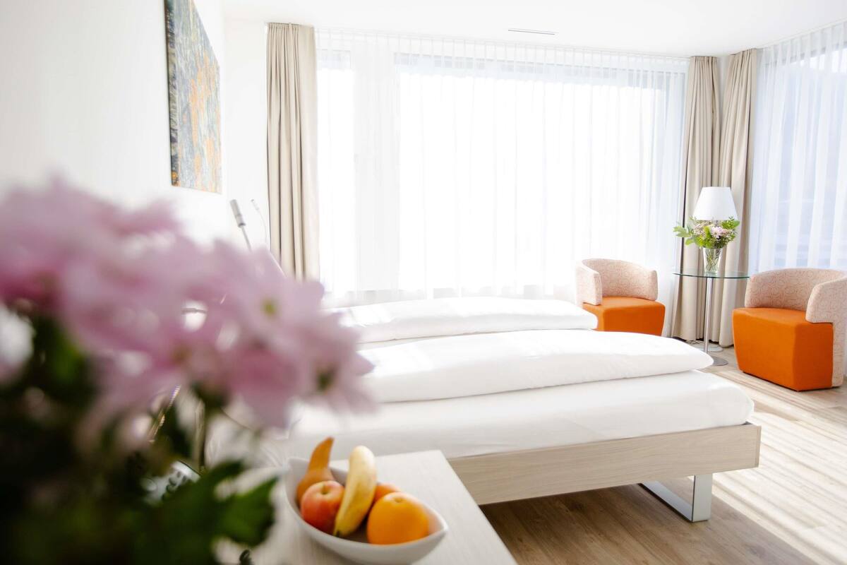 A modern hotel room with a bed, two armchairs, a small table with fruit, and a vase with flowers.