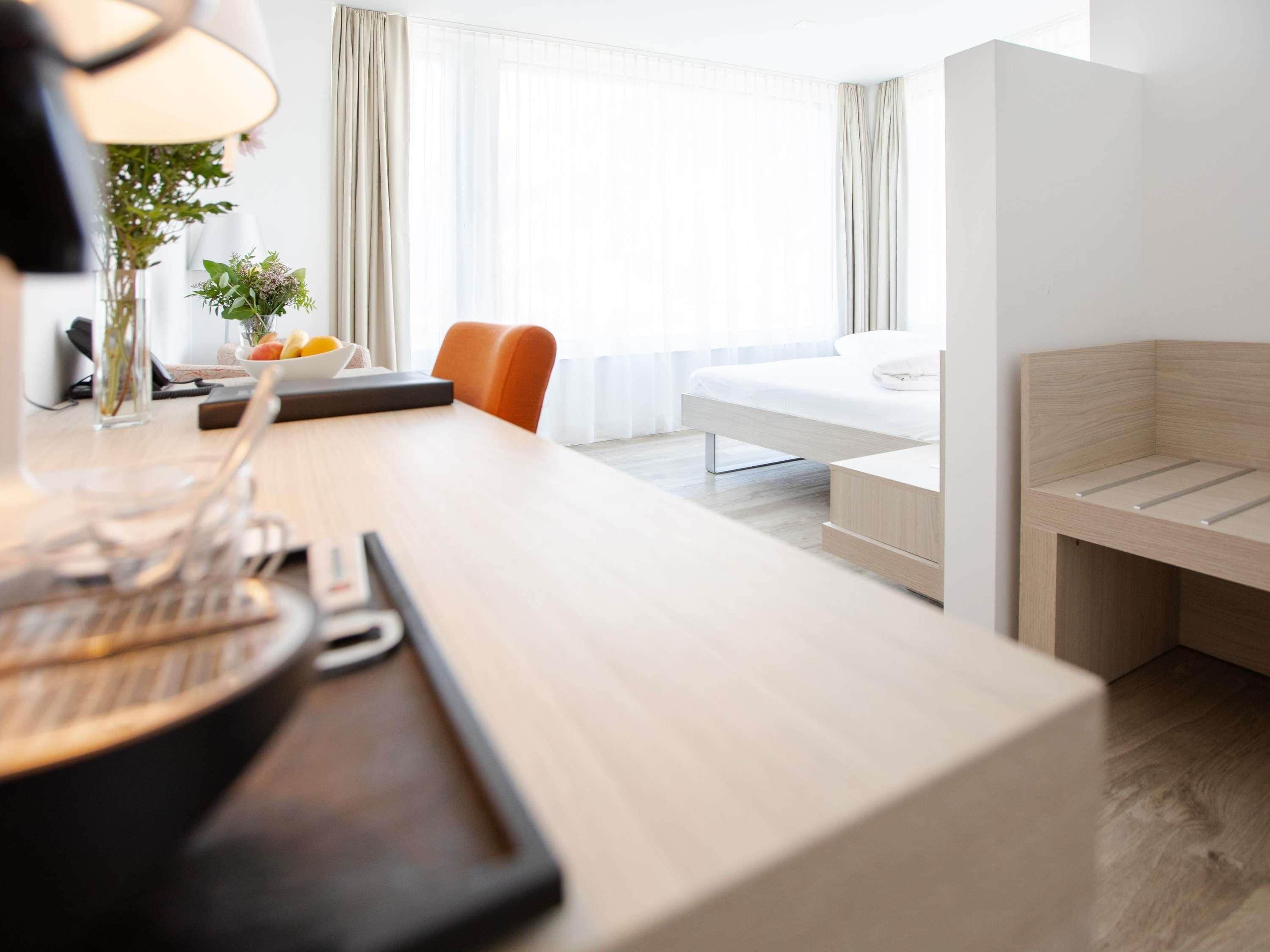 A modern hotel room with a wooden desk, a bed, a chair, and a bedside table.