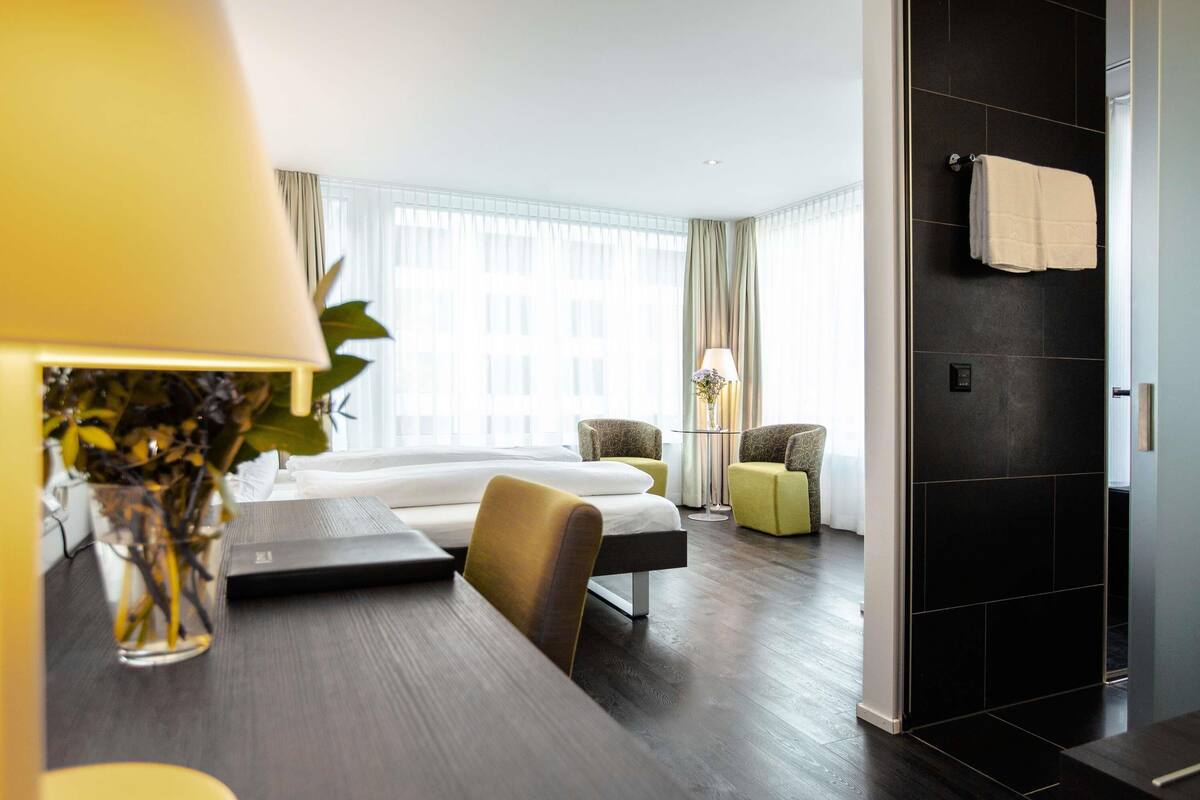 A modern hotel room with a bed, a desk, a chair, and a small table. There is a window with curtains, a lamp, and a plant.