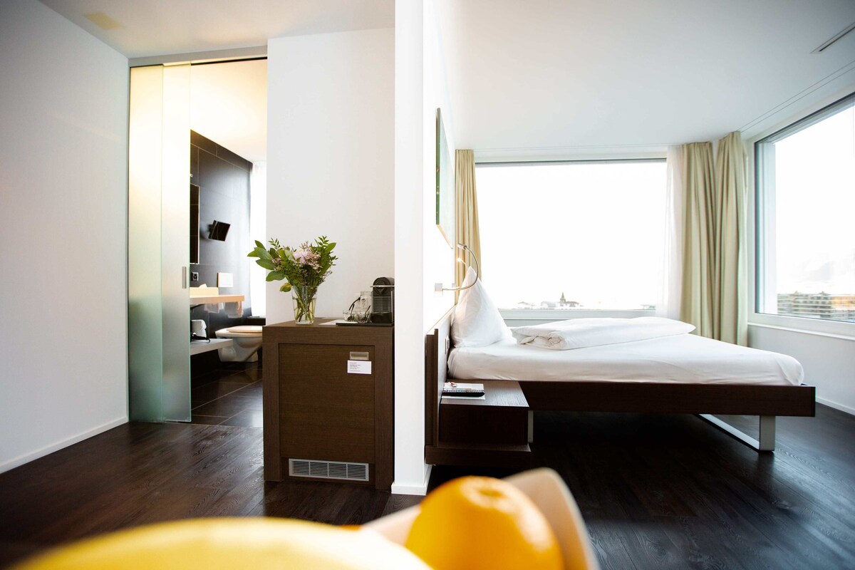 A modern hotel room with a bed, bedside table, and a view of the city.
