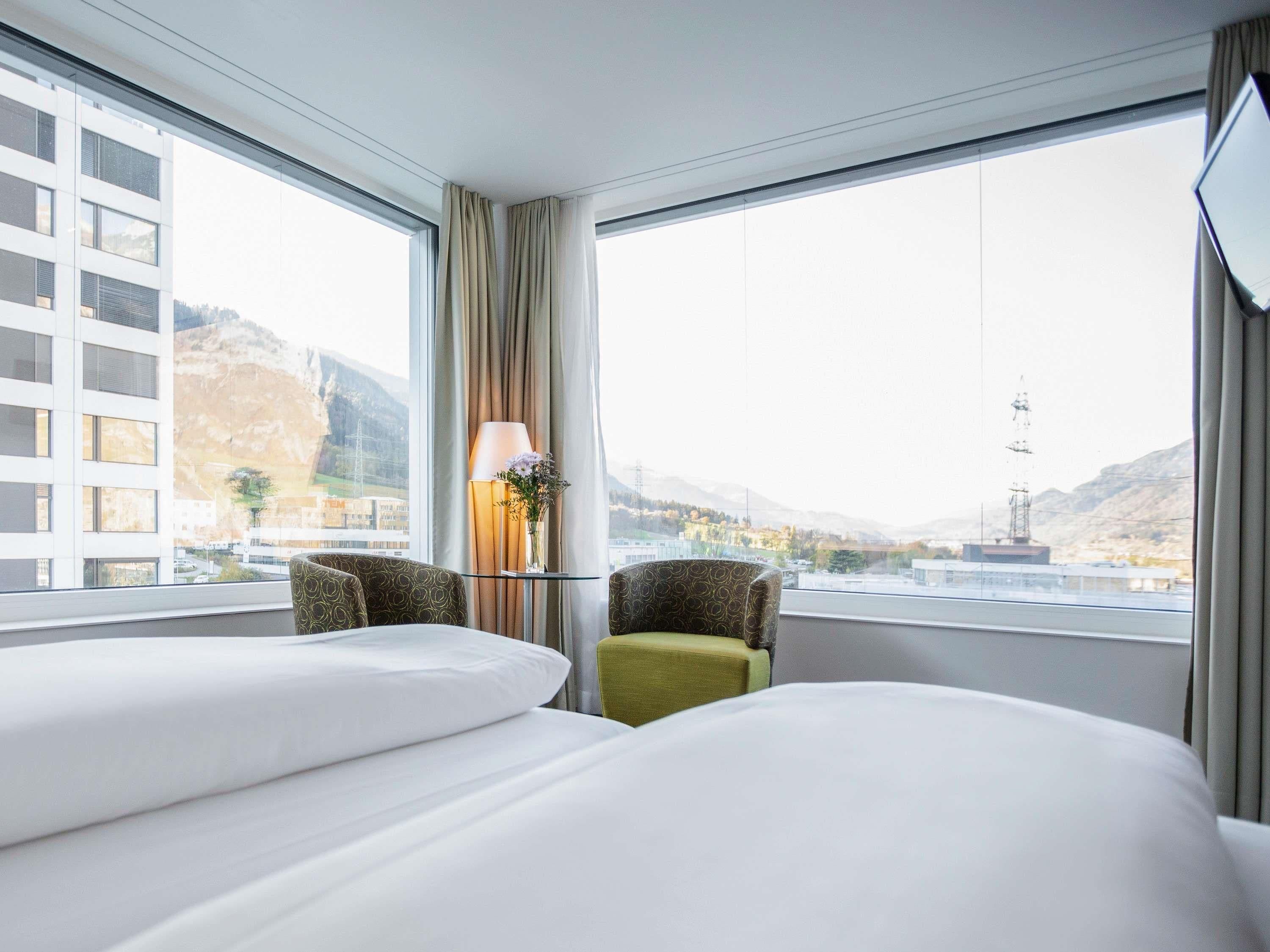 A hotel room with two beds, a lamp, a vase with flowers, and a view of a cityscape and mountains.