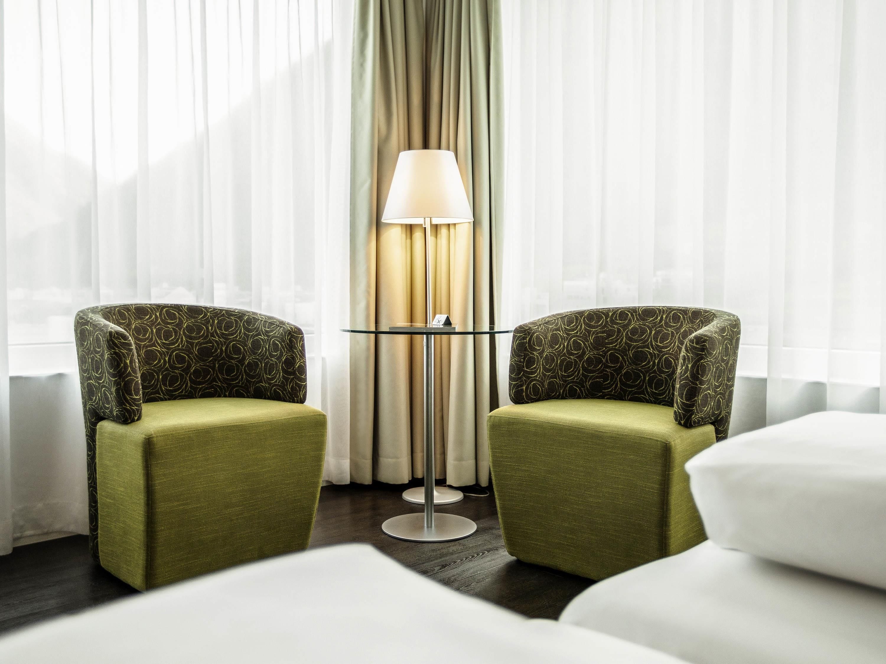 Habitación de hotel con dos sillones verdes, una mesita redonda pequeña, una lámpara y una cama.
