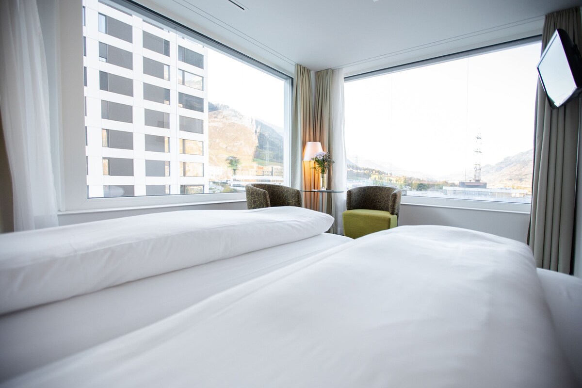 A hotel room with two beds, a desk with a lamp, and large windows offering a view of the cityscape.