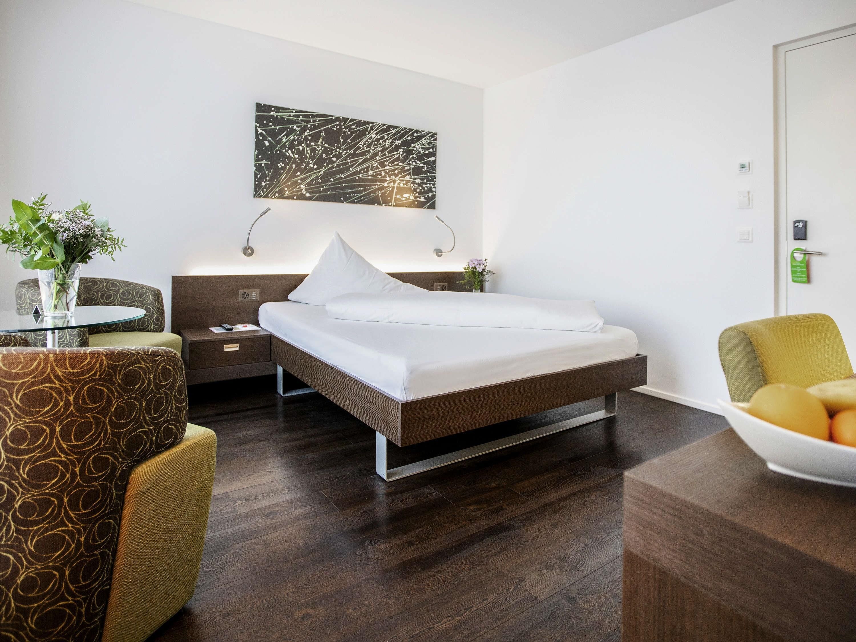A modern hotel room with a bed, a bedside table, a chair, a small table with a vase, and a wall art piece.