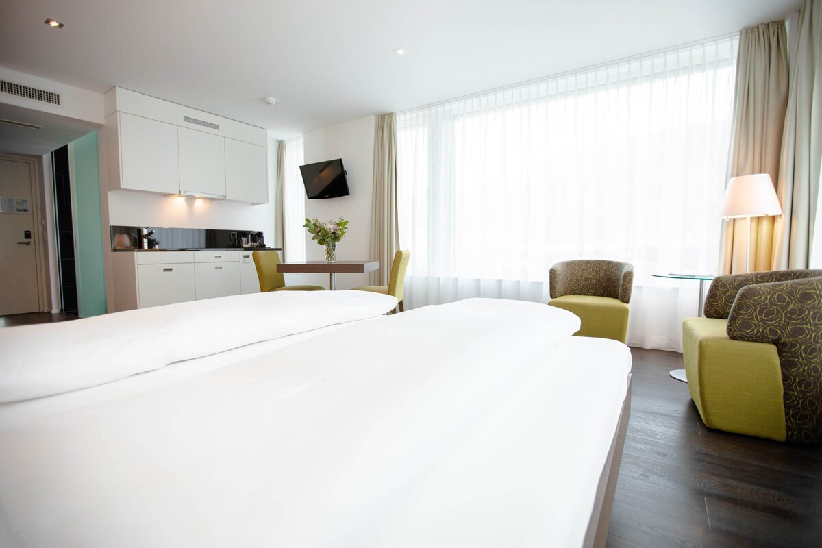 A hotel room with a large bed, a desk with a chair, a small table with a vase, and a TV on the wall.