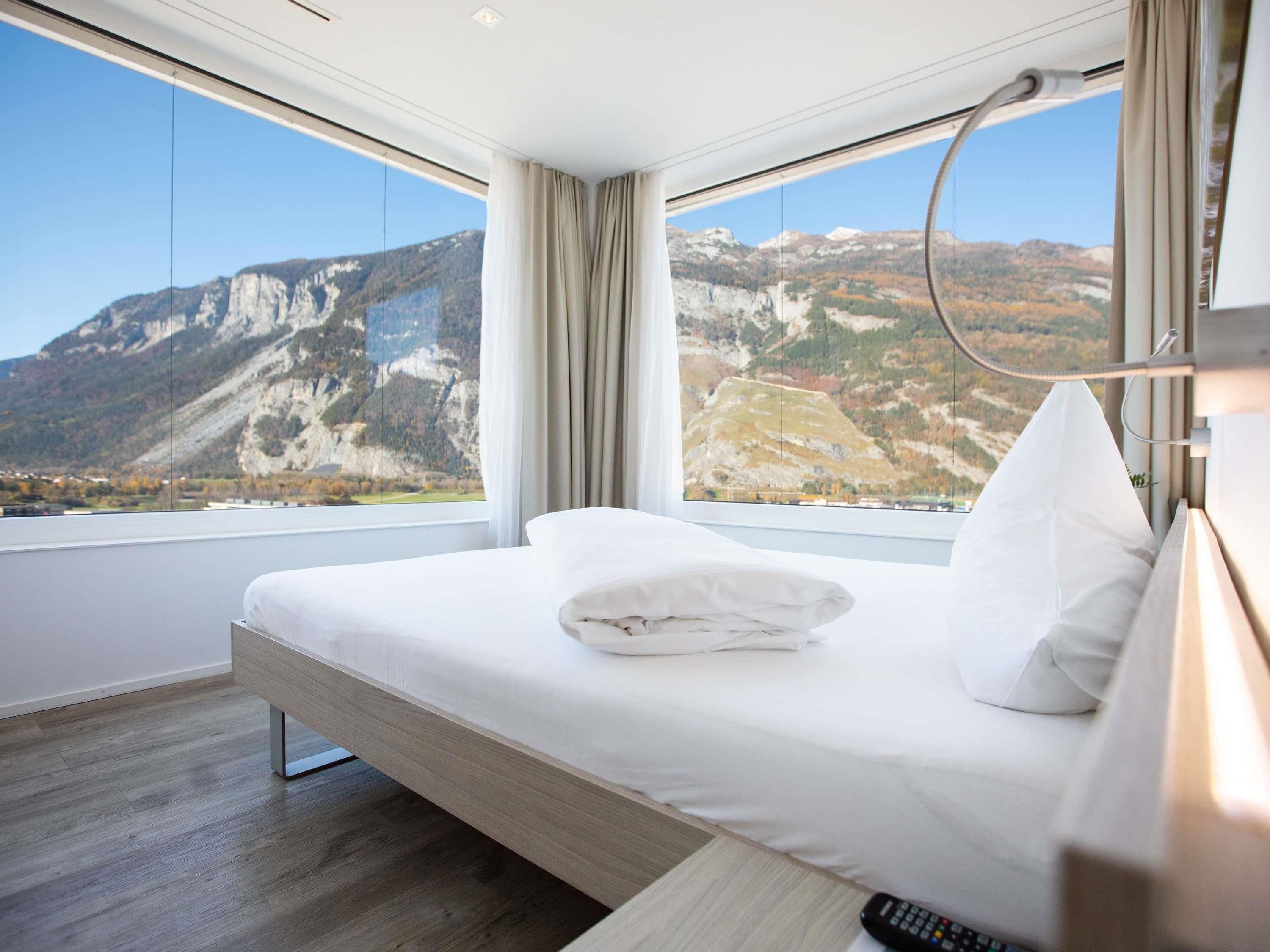 A modern hotel room with a large bed, a remote control, and a view of mountains through floor-to-ceiling windows.