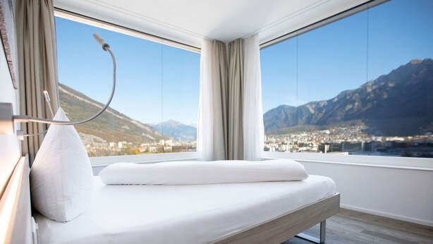 A modern hotel room with a bed, a bedside table, a lamp, and a large window offering a mountain view.