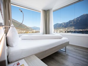 A modern hotel room with a bed, a bedside table, a lamp, and a large window offering a mountain view. - Mercure Chur City West (Chur)