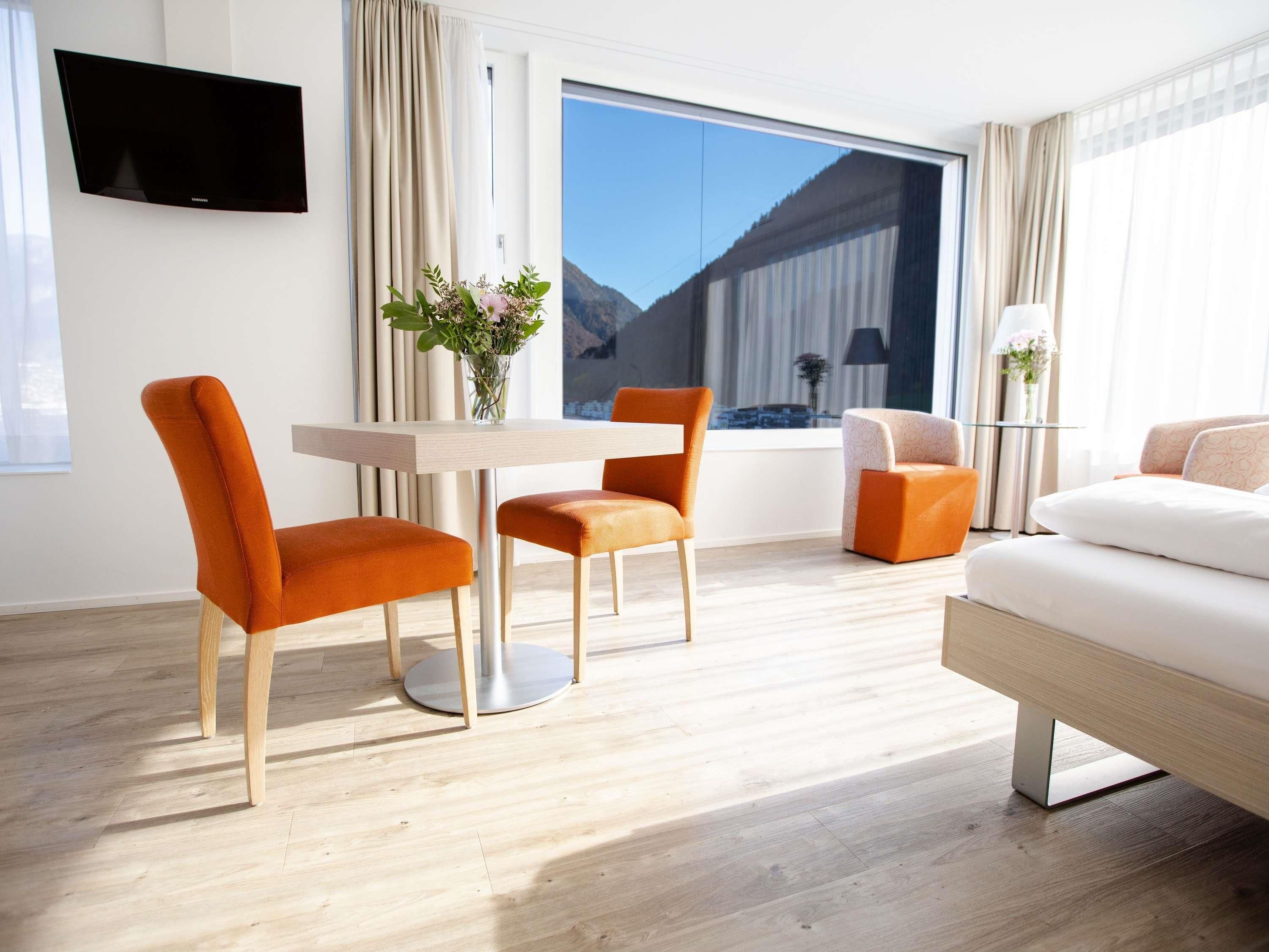 Ein modernes Hotelzimmer mit großem Fenster, einem Flachbildfernseher, einem Esstisch mit zwei Stühlen, einer Couch und einer Lampe.