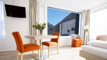 Ein modernes Hotelzimmer mit großem Fenster, einem Flachbildfernseher, einem Esstisch mit zwei Stühlen, einer Couch und einer Lampe.