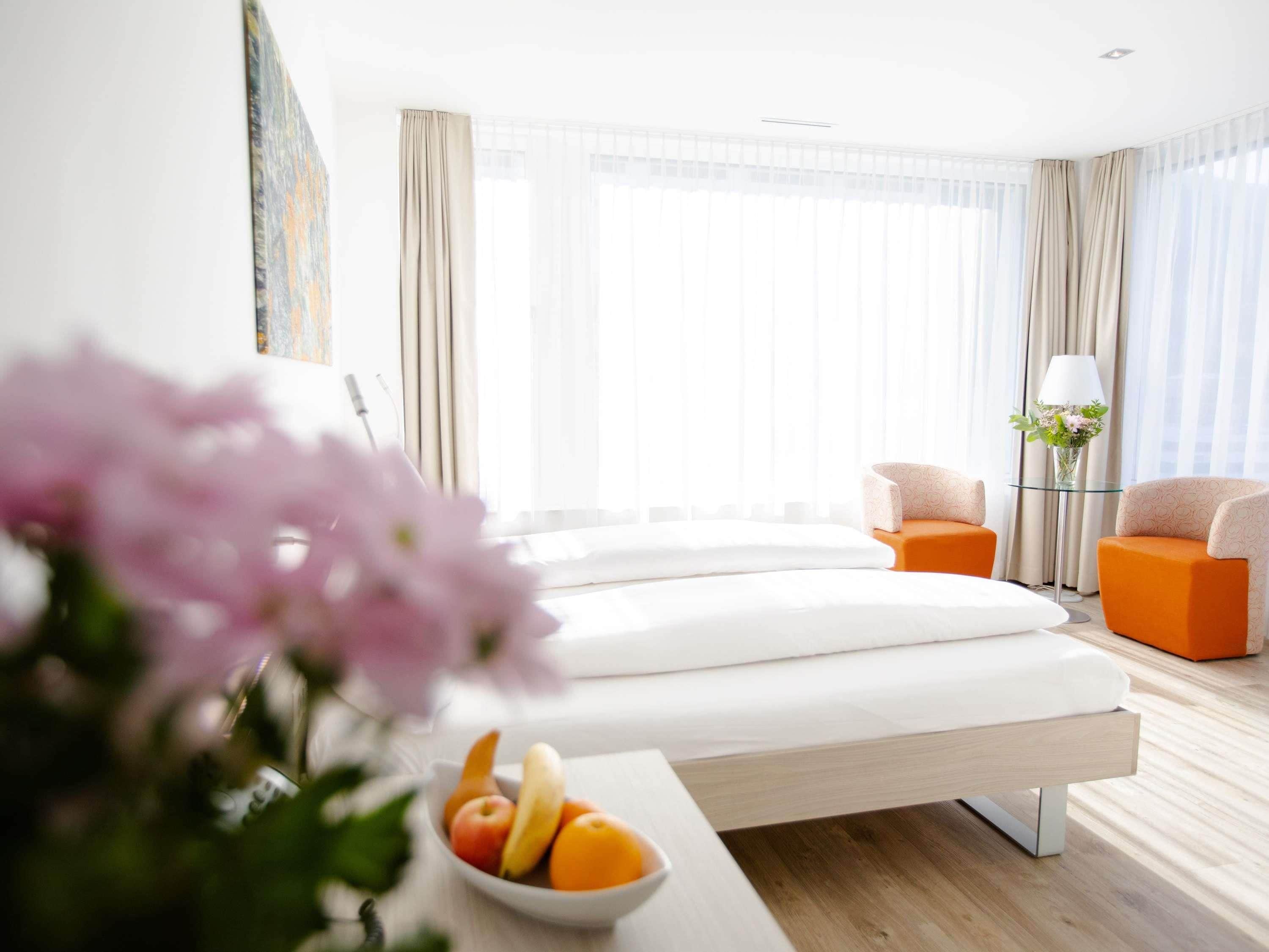 A modern hotel room with a bed, two armchairs, a small table with fruit, and a vase with flowers.