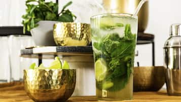Un bicchiere di bevanda ghiacciata con foglie di menta e fette di lime, una ciotola dorata e uno shaker per cocktail su un piano di legno.