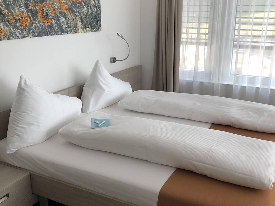 Una cama bien hecha con sábanas blancas y una manta marrón, una mesita de noche con una lámpara y un cuadro en la pared.
