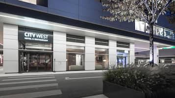 Ein moderner Hotelaufgang mit Glasfassade, einem Schild für das CITY WEST HOTEL & RESTAURANT und einer begrünten Fläche mit einem Baum.