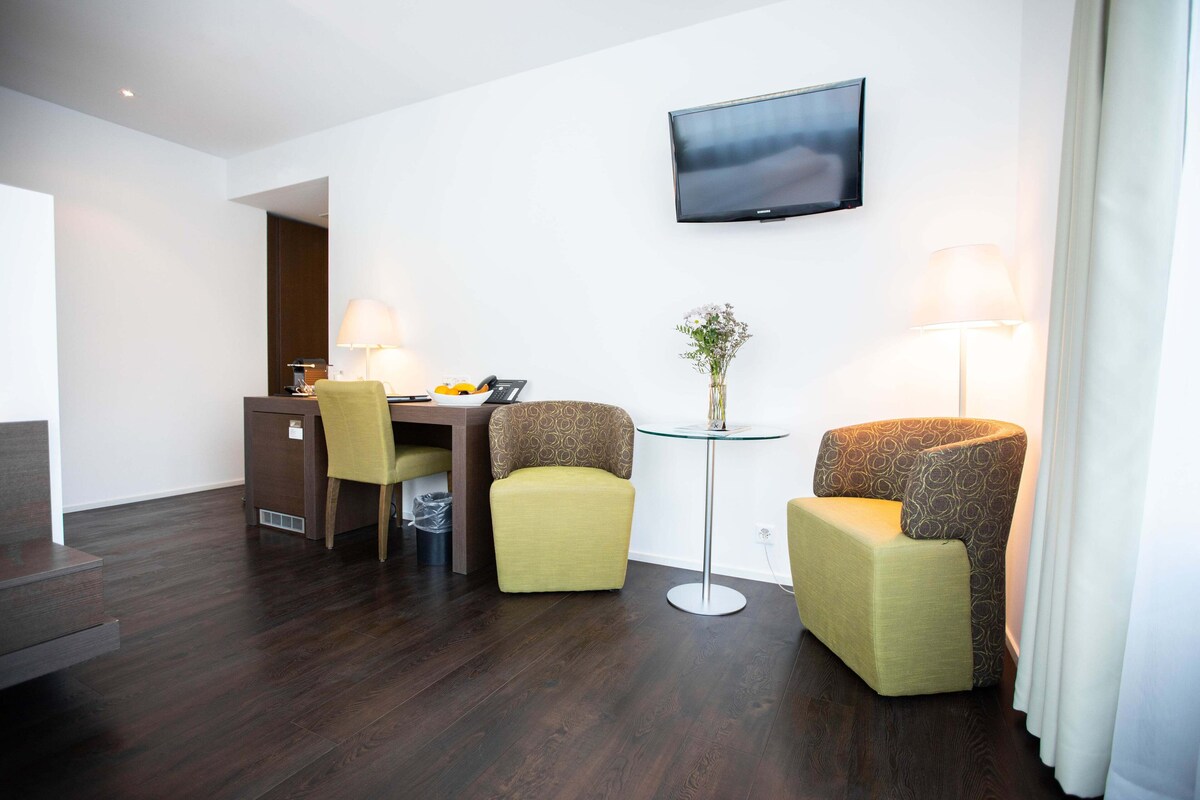 A hotel room with a desk, two armchairs, a TV mounted on the wall, and a small table with a vase of flowers.