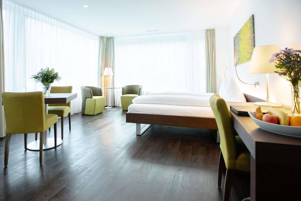 A hotel room with a bed, two chairs, a table, and a desk with a fruit bowl.