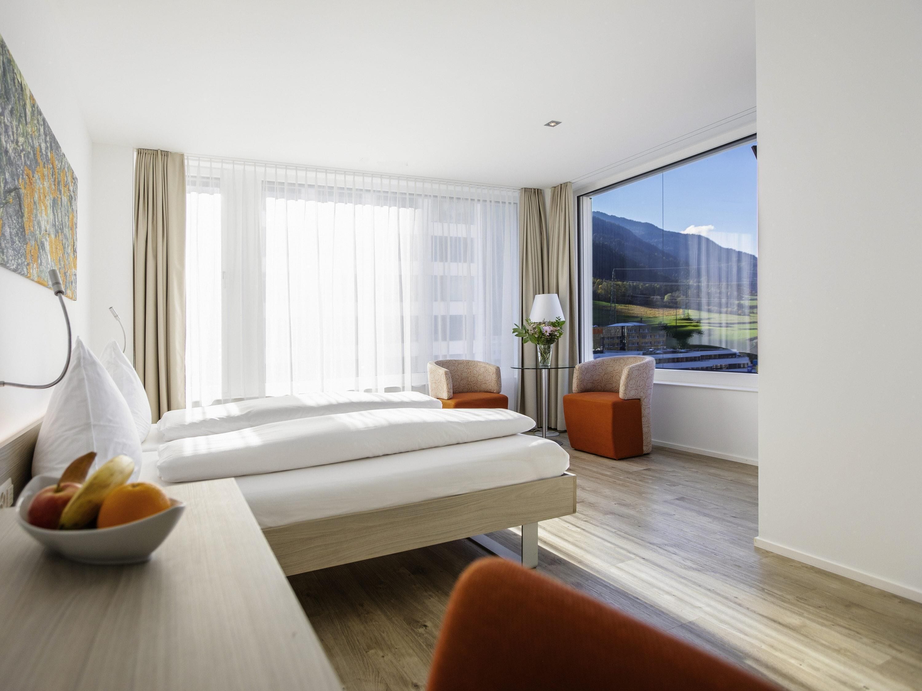 A hotel room with a bed, two chairs, a table with fruit, a lamp, and a view of mountains.