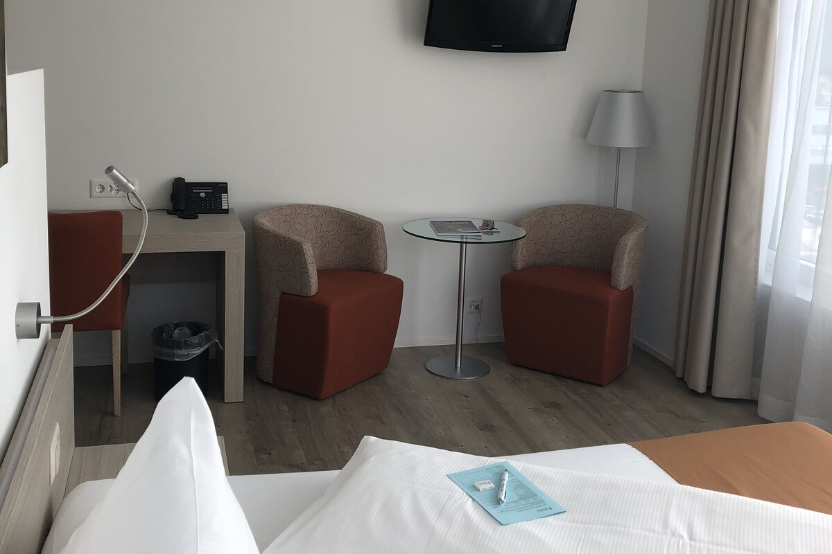 A hotel room with a bed, two armchairs, a small table, a TV mounted on the wall, a lamp, and a window with curtains.
