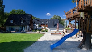 A playground with a blue slide and wooden climbing structures, set against a house with a sloped roof and a gravel path leading to it. - Apartmán-Hotel Jítrava (Rynoltice)