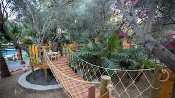 Un puente de madera sobre una zona arenosa con una red de cuerda, rodeado de árboles y plantas.