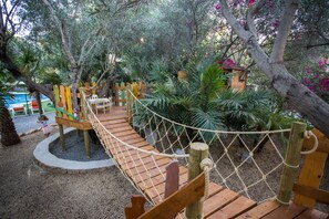 Un pont en bois enjambant une zone sablonneuse, muni d’un filet de corde, et entouré d’arbres et de plantes.