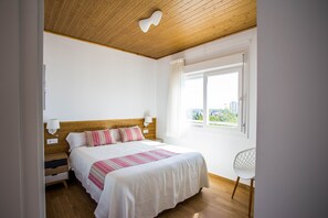 Een slaapkamer met een houten plafond, een groot bed met een gestreept dekbed, twee nachtkastjes met lampen, een witte stoel en een raam met uitzicht op gebouwen.