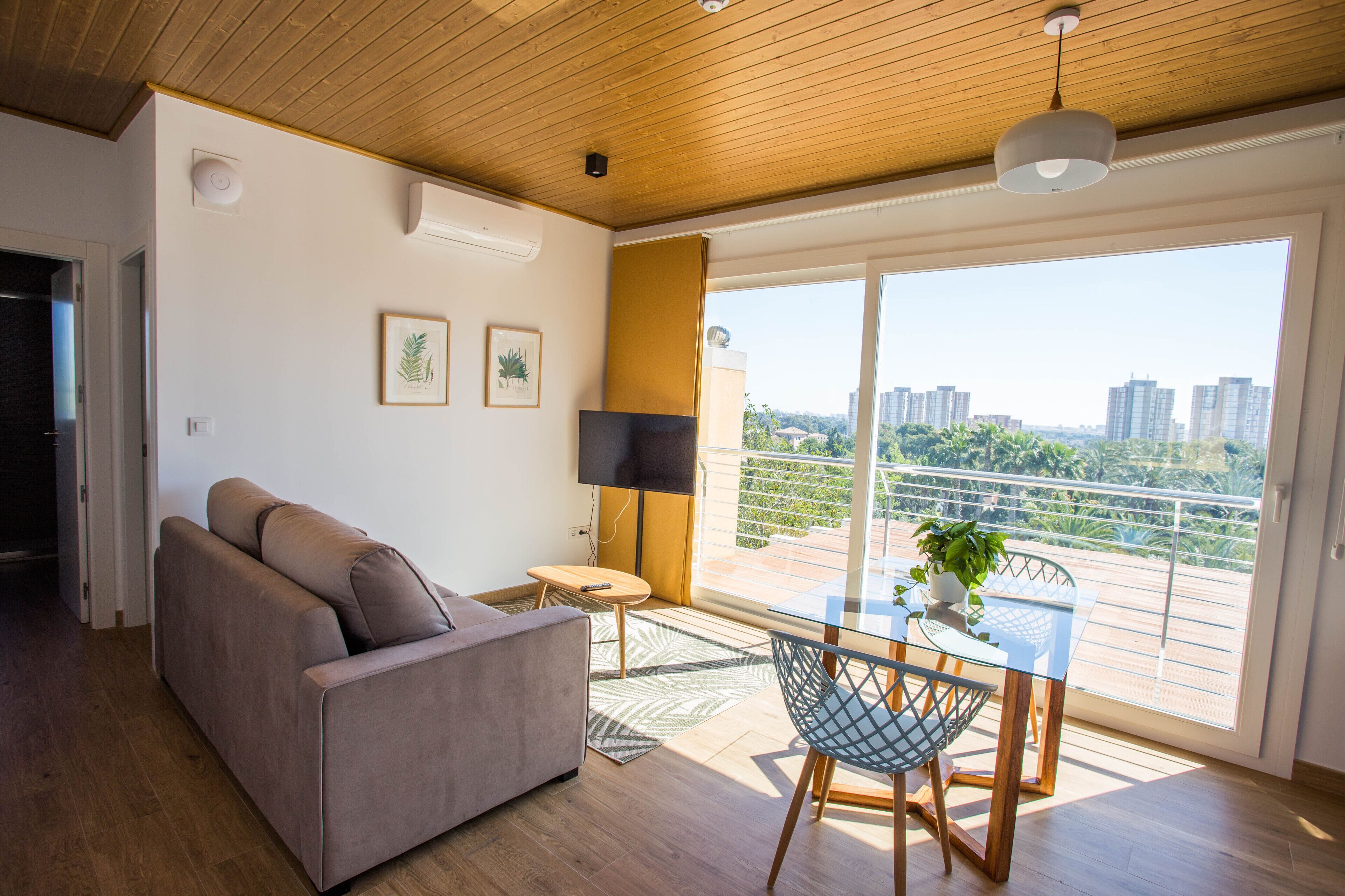 A modern living room with a sofa, a small table with a plant, and a balcony with a view of buildings.