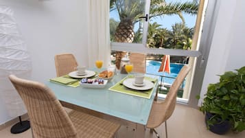 Comedor con mesa de vidrio, sillas de mimbre, alimentos para el desayuno y vista a una piscina y palmeras.