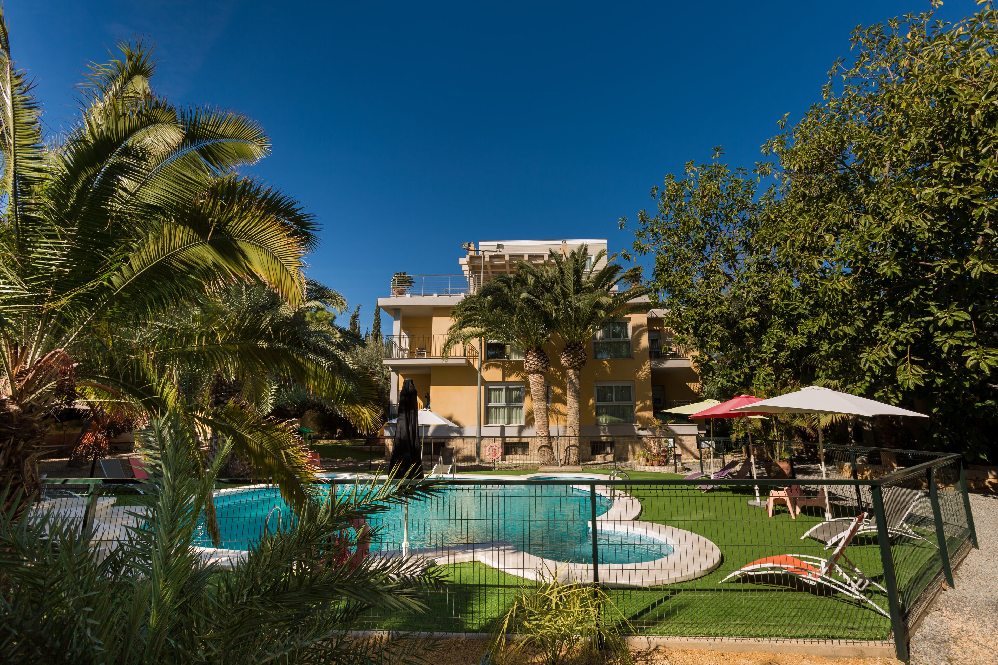 A swimming pool with a green deck, surrounded by palm trees and a building with balconies.