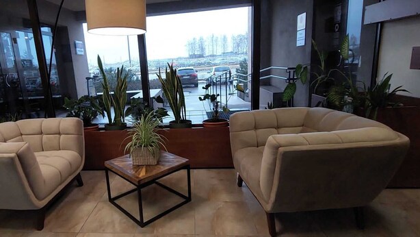 Un área de descanso moderna con dos sillones color beige, una mesa de centro de madera con una planta en maceta, y ventanas amplias con vista a un estacionamiento.