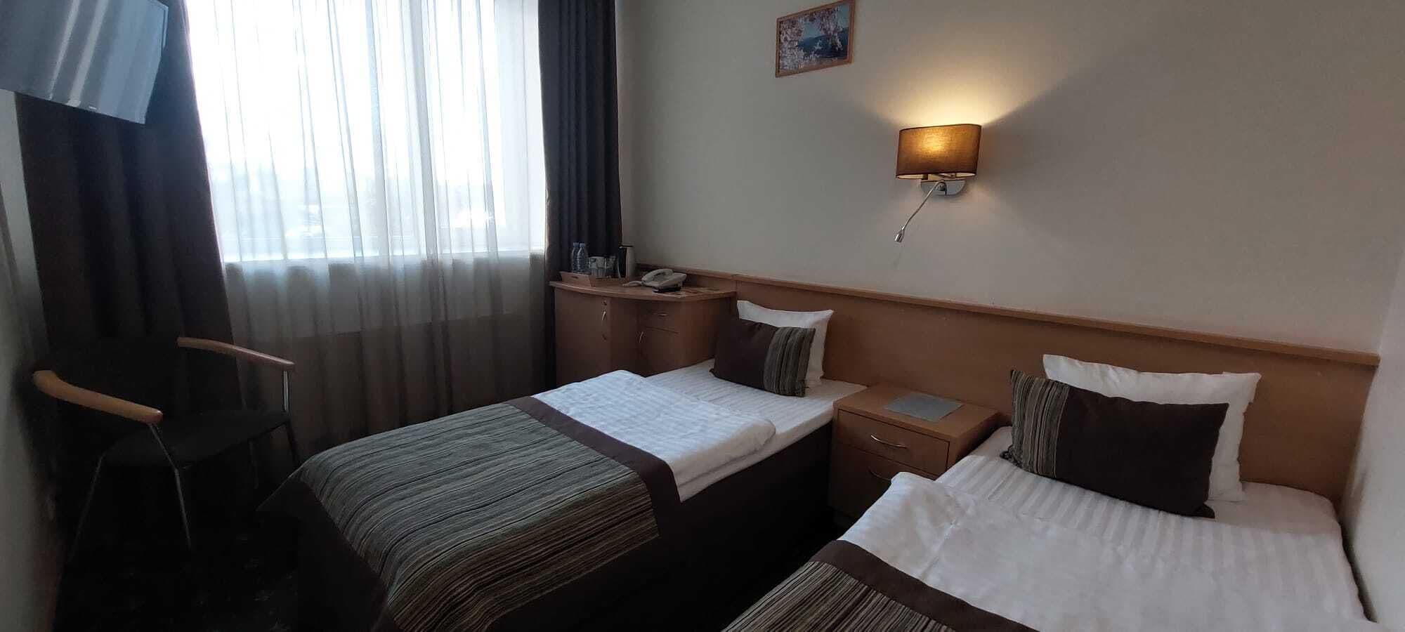 A hotel room with two single beds, a desk, a chair, a bedside table, and a wall-mounted lamp.