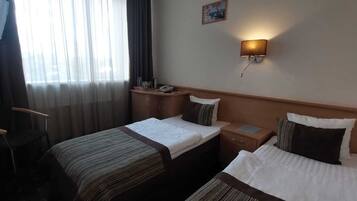 A hotel room with two single beds, a desk, a chair, a bedside table, and a wall-mounted lamp.