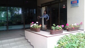 Entrada de un hotel con una mujer de pie junto a la puerta, un cartel que dice "SKY HIKHI VIESnica Hotel" y tres cestas de mimbre con flores en los escalones.