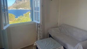 Ett rum med ett fönster som vetter mot en sjö eller ett hav och ett berg, en vit soffa och ett litet bord med ett blommigt bordsduk.