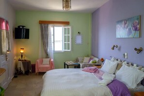 A bedroom with a bed, a chair, a table, a TV, and a window with curtains. - Melina's sunset (Kalymnos)