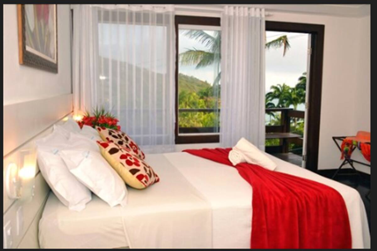 Une chambre d’hôtel avec un lit, du linge de lit blanc, un plaid rouge, un oreiller à motifs rouges et blancs, une fenêtre donnant sur un espace verdoyant et une porte menant à un espace extérieur.