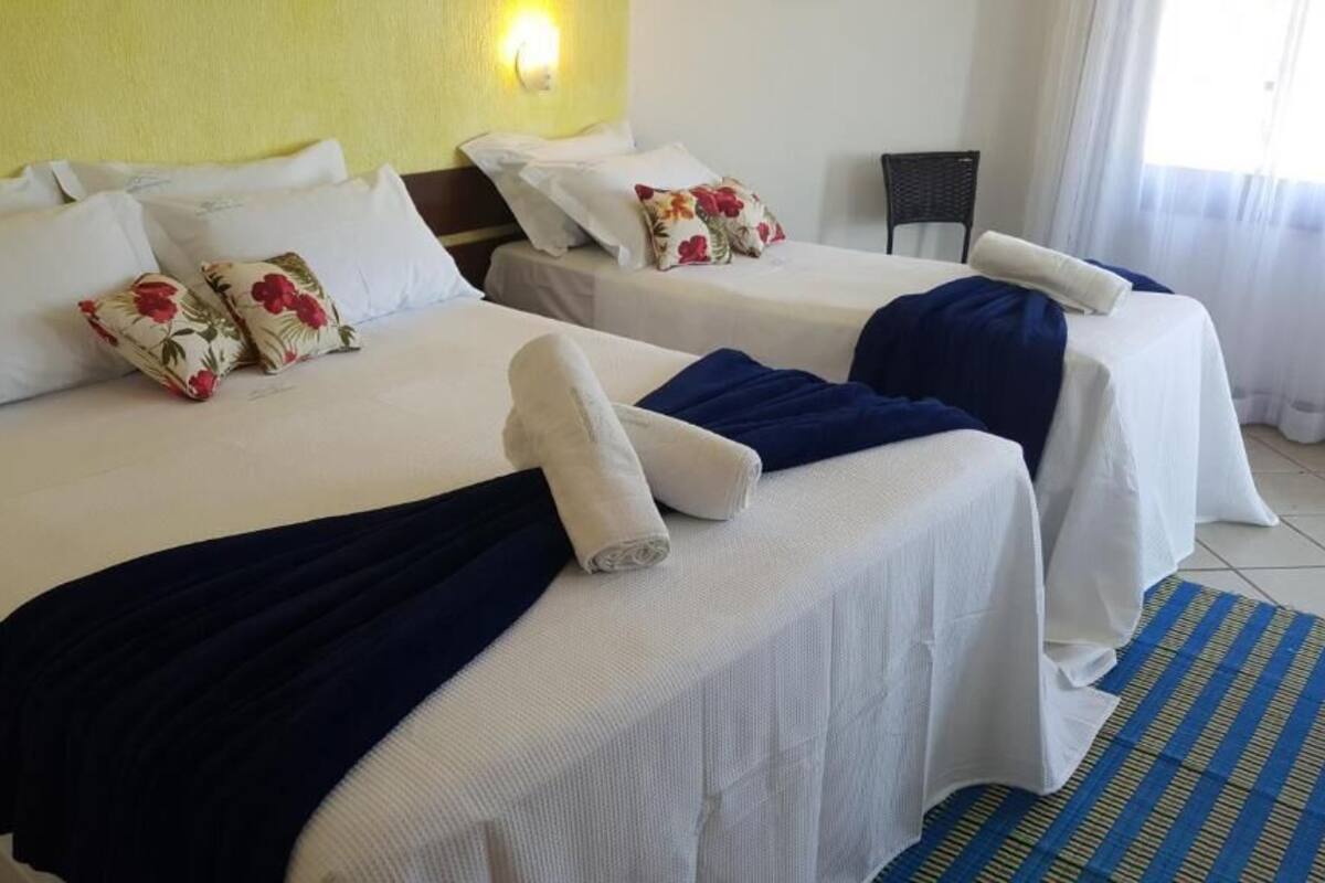 Une chambre d’hôtel avec deux lits, du linge de lit blanc, des coussins à motifs floraux, une couverture bleue, un tapis à rayures et une chaise.