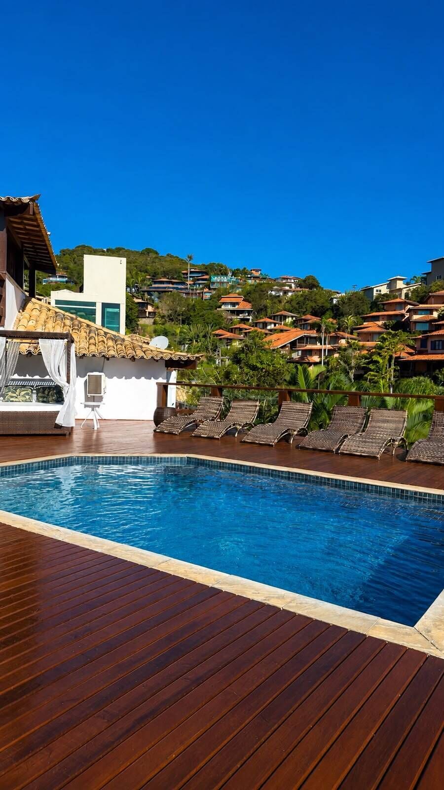 A swimming pool with wooden deck, lounge chairs, and a view of hillside houses.