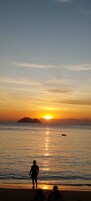 Escena de playa con atardecer, agua tranquila y siluetas de personas.