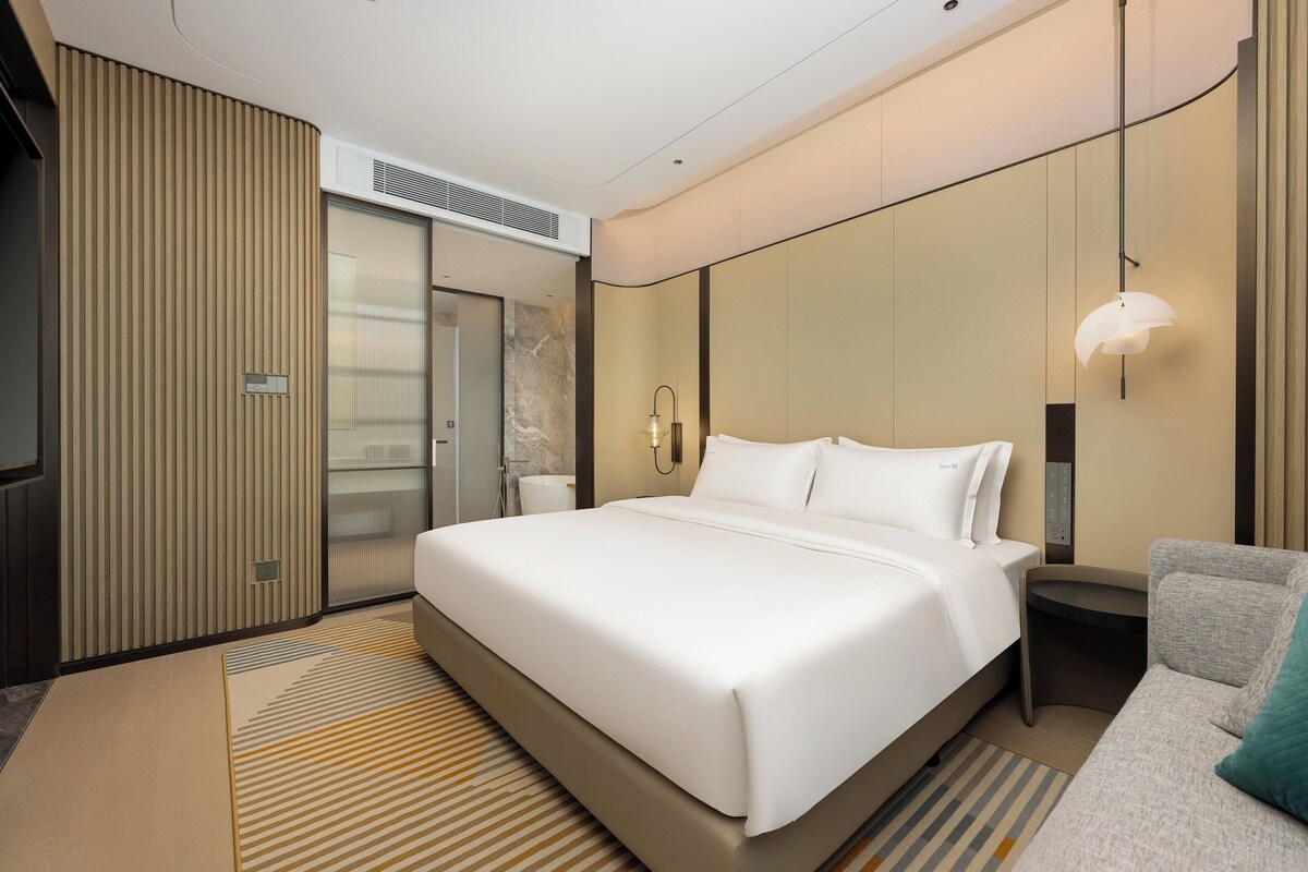 Una habitación de hotel moderna con una cama grande, una mesita de noche, un sofá, un televisor montado en la pared y un baño con ducha e inodoro.