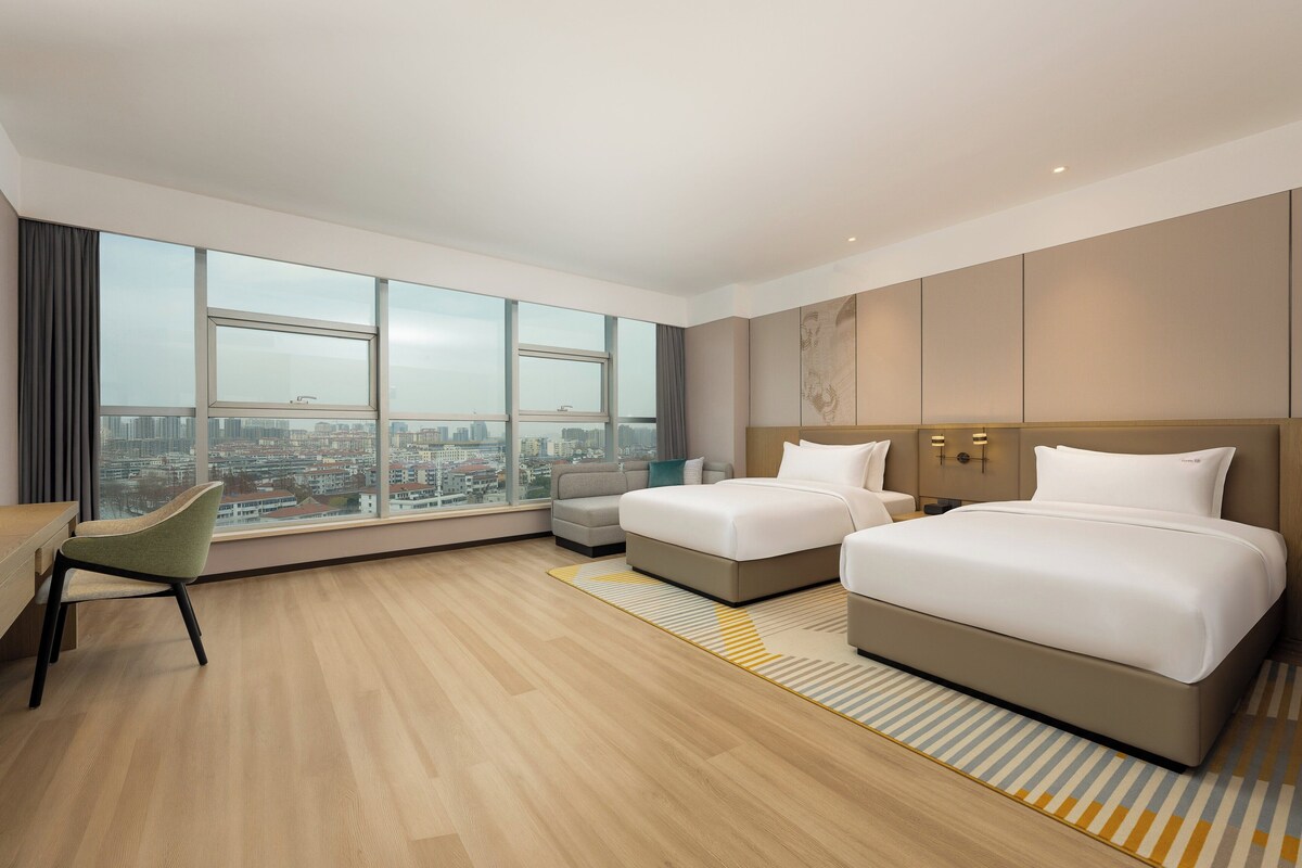 Habitación de hotel moderna con dos camas, un escritorio y un ventanal grande con vistas a la ciudad.