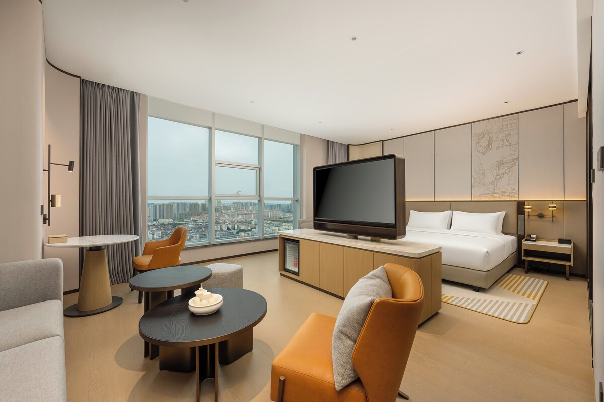 Habitación de hotel moderna con televisor grande, asientos cómodos, una cama y vistas a la ciudad.